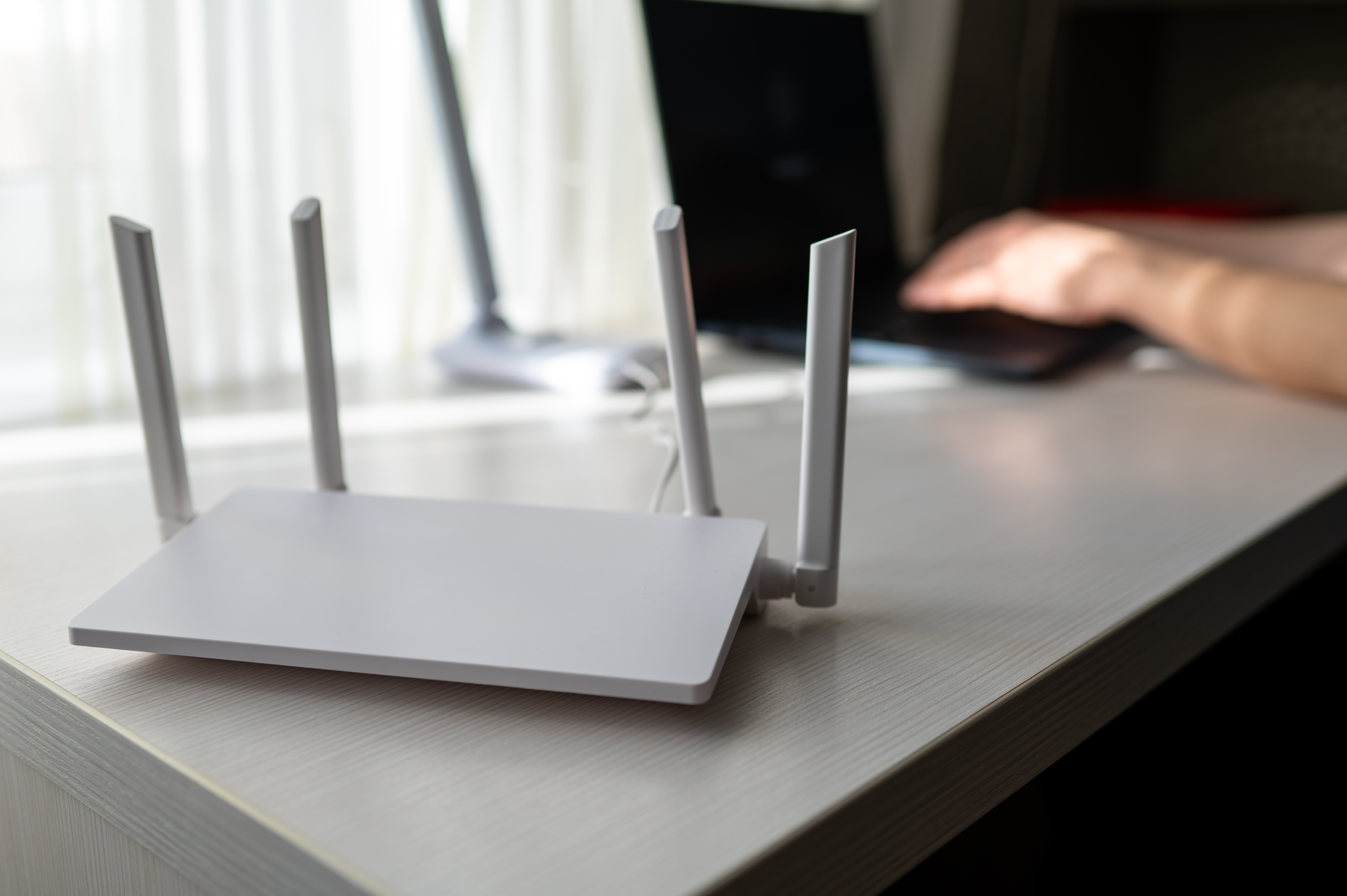 Close-up of a modern wireless router with antennas on a wooden desk. In the blurred background, a young woman is working on a laptop, representing high-speed internet and remote work lifestyle
