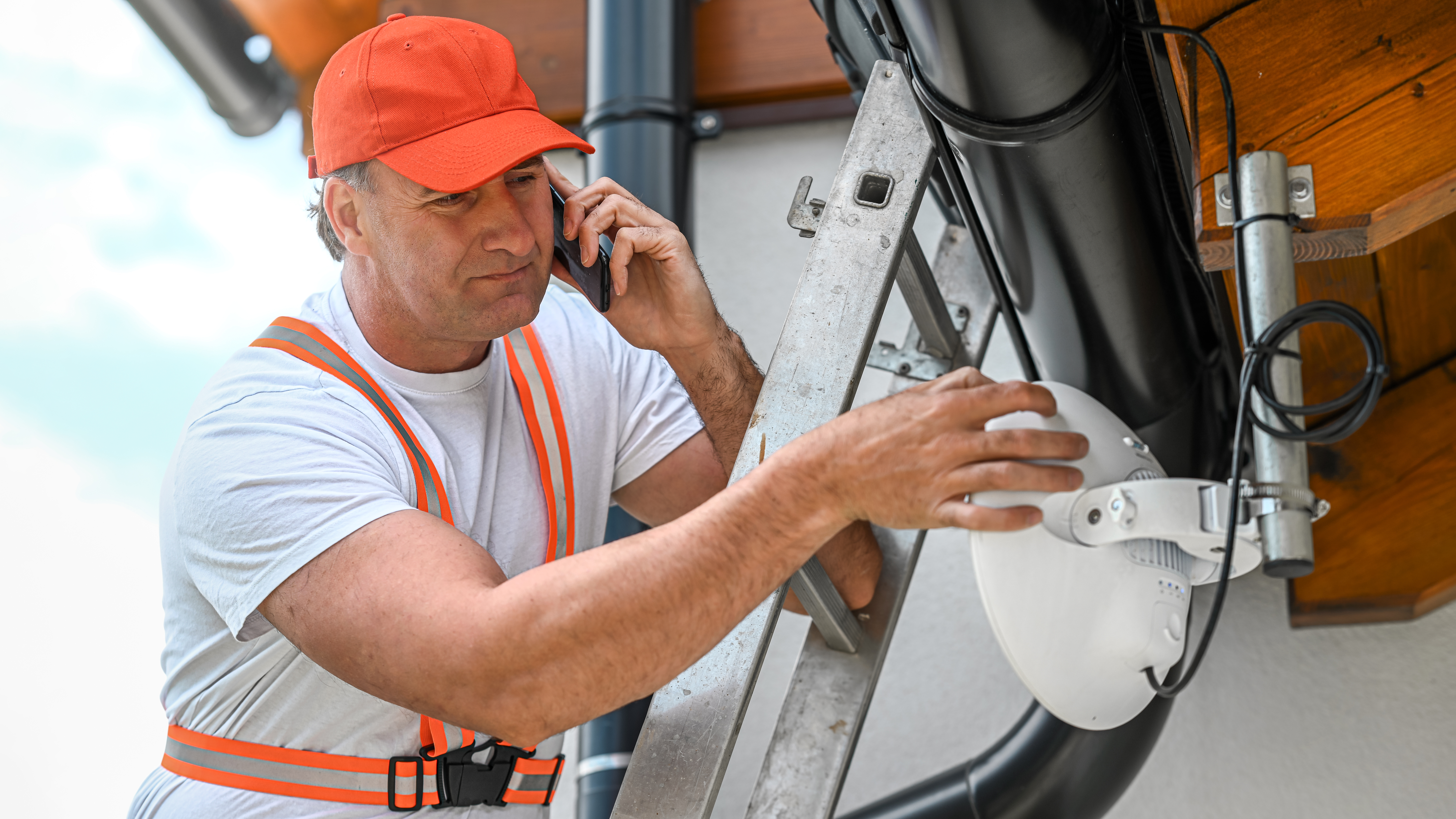 Technician on aluminium ladder communicates with a colleague by phoneduring to adjustment of directional WiFi antenna. Network infrastructure maintenance concept.